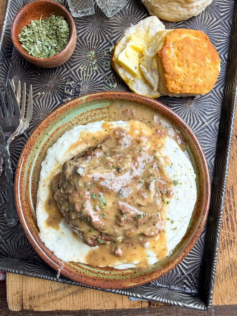 A plate of mashed potatoes topped with brown gravy and herbs, served alongside a buttered biscuit and a small bowl of dried parsley on a textured tray with two forks.