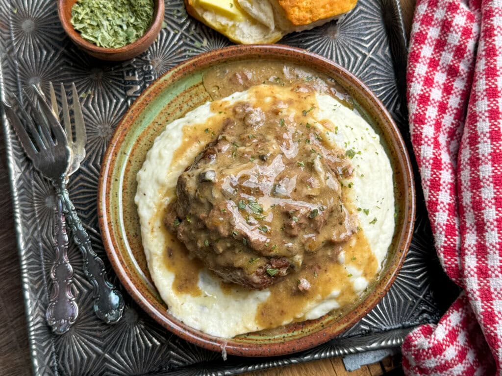A plate with mashed potatoes topped with a hamburger patty and brown gravy, garnished with herbs. A biscuit with butter, a fork and spoon, a small bowl of dried herbs, and a red checked cloth are beside the plate.