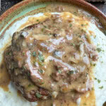 A close-up of poor man's steak topped with brown gravy, served on a bed of creamy mashed potatoes. A sprinkle of herbs is visible on top, with a biscuit and a butter in the background.