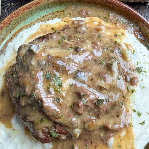 A close-up of poor man's steak topped with brown gravy, served on a bed of creamy mashed potatoes. A sprinkle of herbs is visible on top, with a biscuit and a butter in the background.