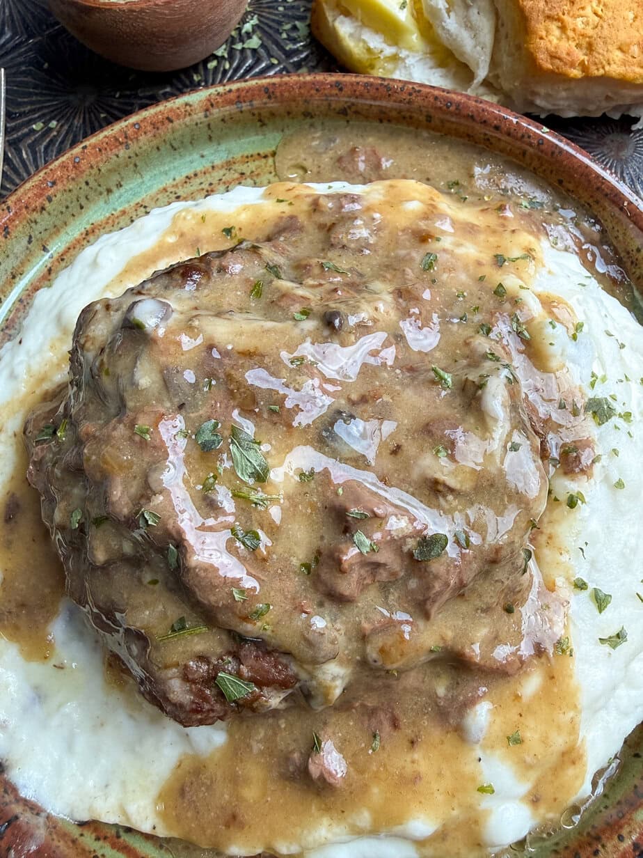 A close-up of poor man's steak topped with brown gravy, served on a bed of creamy mashed potatoes. A sprinkle of herbs is visible on top, with a biscuit and a butter in the background.