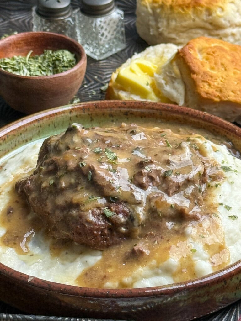 A bowl of mashed potatoes topped with a piece of beef and brown gravy, sprinkled with herbs. In the background are flaky biscuits with butter, a bowl of dried herbs, and salt and pepper shakers.