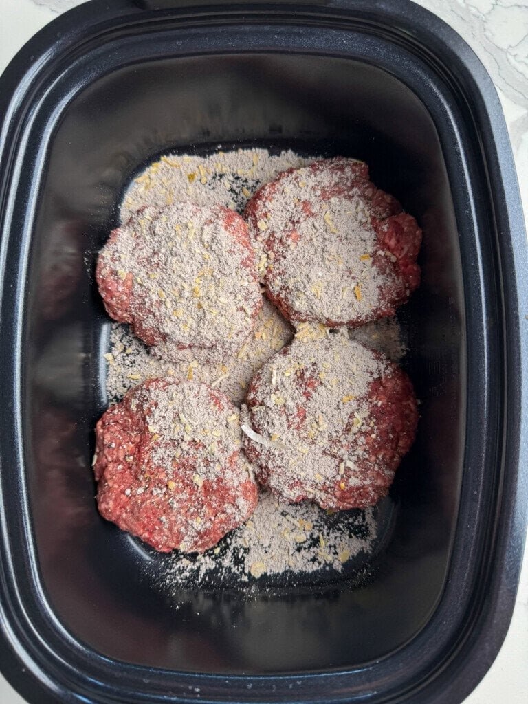 Four raw beef patties covered with dry onion soup mix sit in a black slow cooker, ready to be cooked. The patties are arranged in a single layer on the bottom of the cooker.
