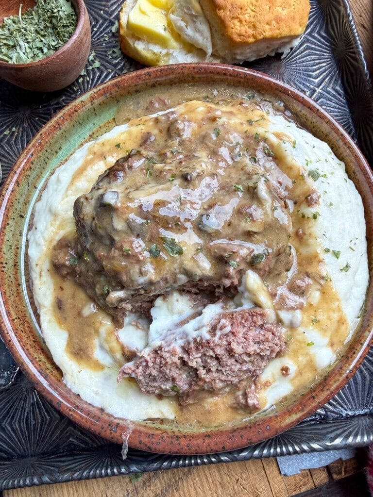 A plate of mashed potatoes topped with a slice of beef covered in brown gravy, garnished with herbs. A biscuit with butter is on the side of the plate.