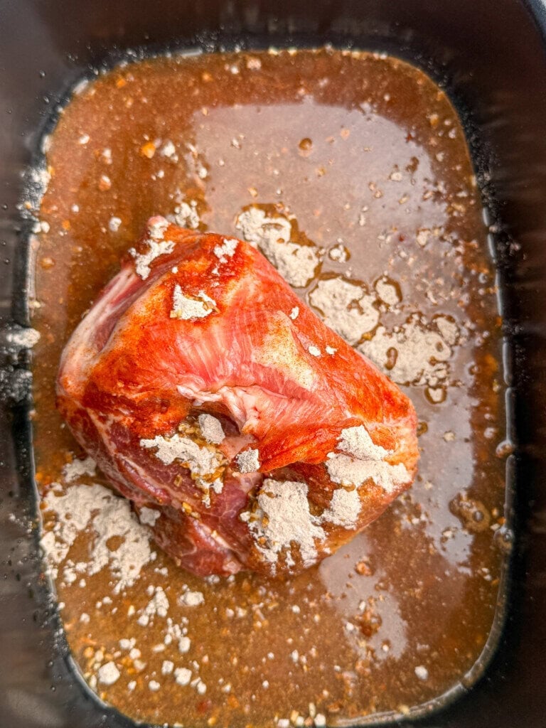 A raw piece of seasoned pork sits in a dark slow cooker, surrounded by a brown marinade with visible seasoning and spice powder on top and in the liquid.