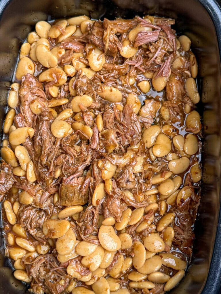 A close-up view of cooked lima beans mixed with shredded meat in a rich brown sauce, all contained in a black slow cooker.