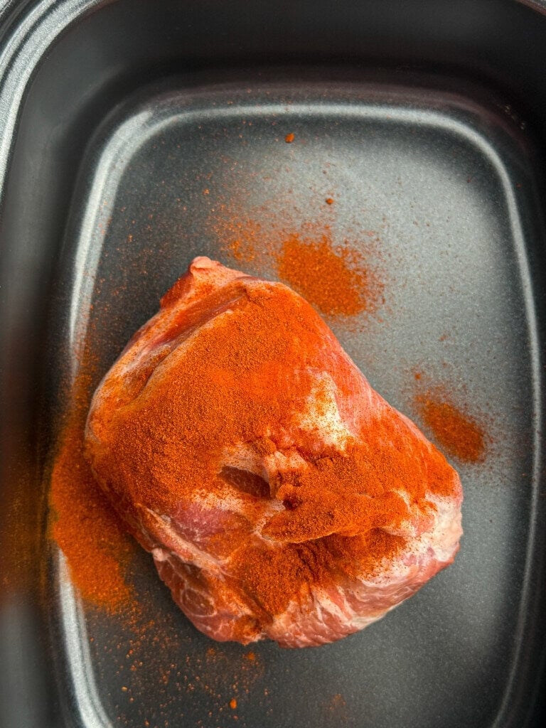 A raw piece of meat coated with paprika, sits in a black slow cooker pan. Some of the red seasoning is sprinkled around the meat inside the pan.