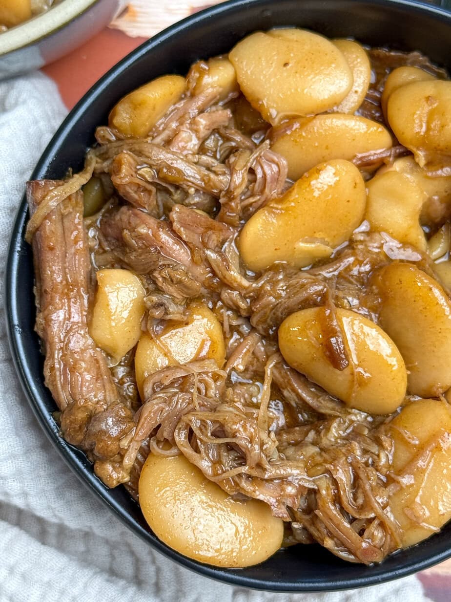 A close-up of a bowl filled with shredded pork and large butter beans in a thick, brown sauce. The dish sits next to a light-colored textured cloth.