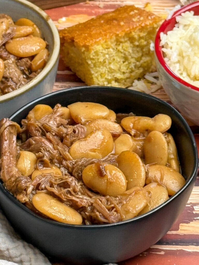 A bowl filled with cooked butter beans and shredded pork butt, accompanied by a slice of cornbread and a bowl of white rice in the background.