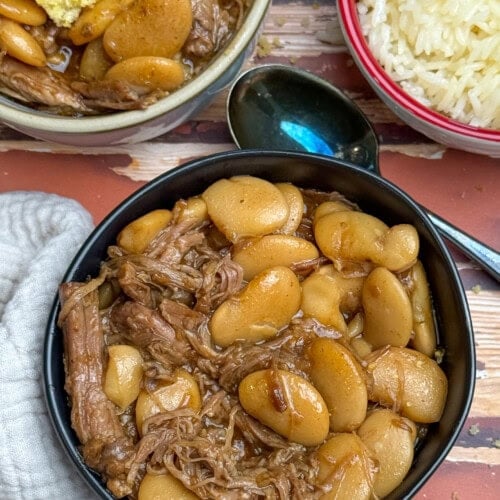 A bowl of cooked butter beans and shredded pork butt in a savory brown sauce, with a spoon beside it. Nearby are a bowl of fluffy white rice and a dish of cornbread on a rustic wooden table.