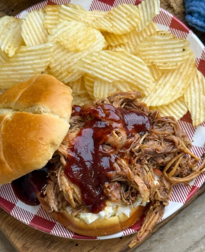 A plate with a pulled pork sandwich topped with barbecue sauce and served with wavy potato chips on a red and white checkered paper.