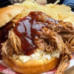 A pulled pork sandwich topped with barbecue sauce and coleslaw is served on a plate with a side of ridged potato chips. A cup of barbecue sauce and a cup of coleslaw are in the background.