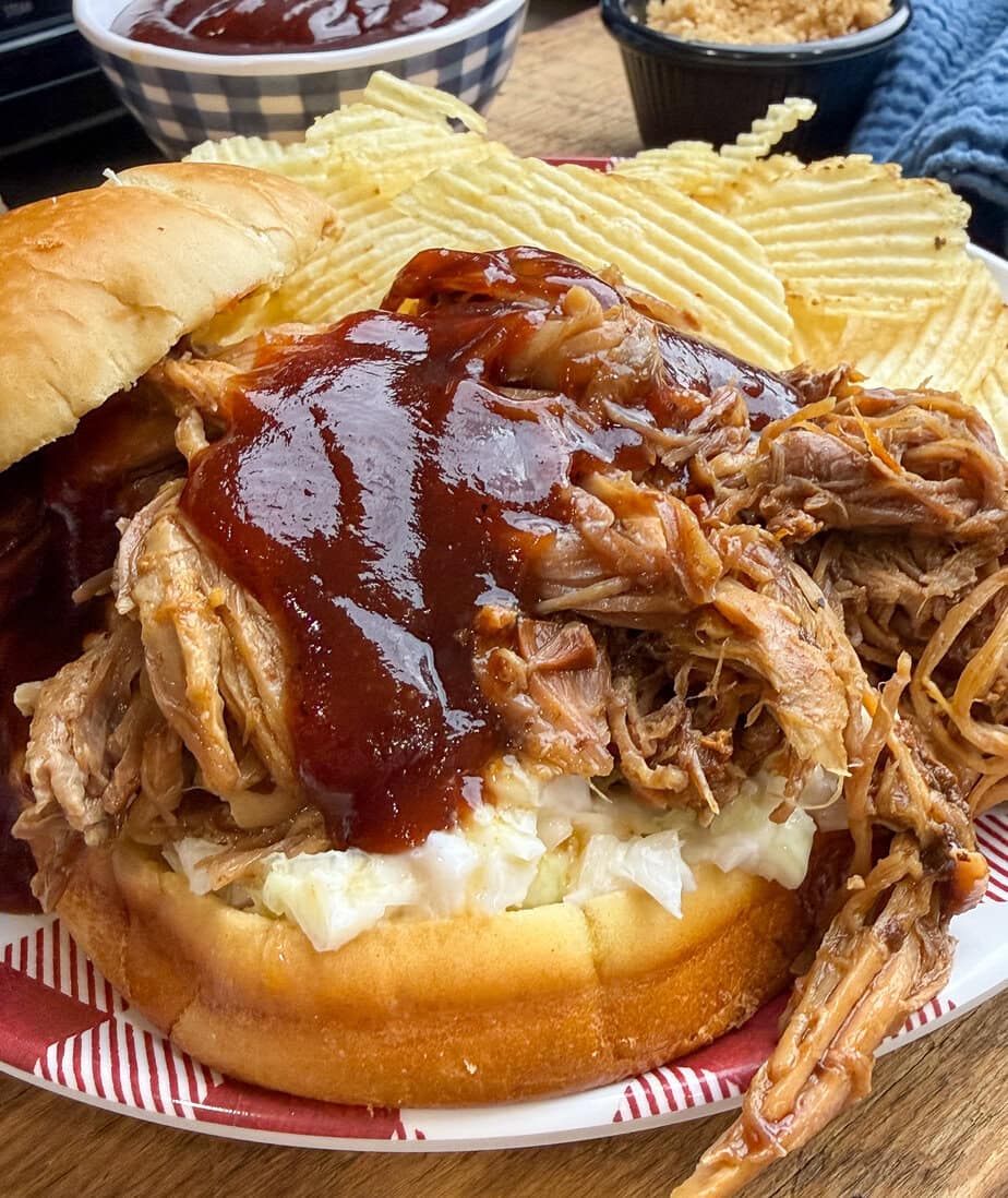 A pulled pork sandwich topped with barbecue sauce and coleslaw is served on a plate with a side of ridged potato chips. A cup of barbecue sauce and a cup of coleslaw are in the background.