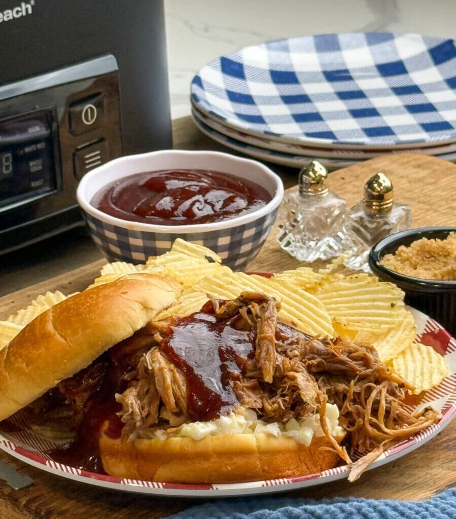 A pulled pork sandwich with barbecue sauce sits on a plate with potato chips. In the background are a bowl of barbecue sauce, a plate, a slow cooker, salt and pepper shakers, and a small dish of brown sugar.