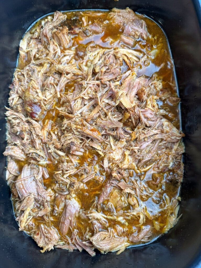 Shredded pulled pork in a rich, brown sauce, filling a rectangular slow cooker or roasting pan. The meat appears tender and juicy, mixed with the flavorful liquid.