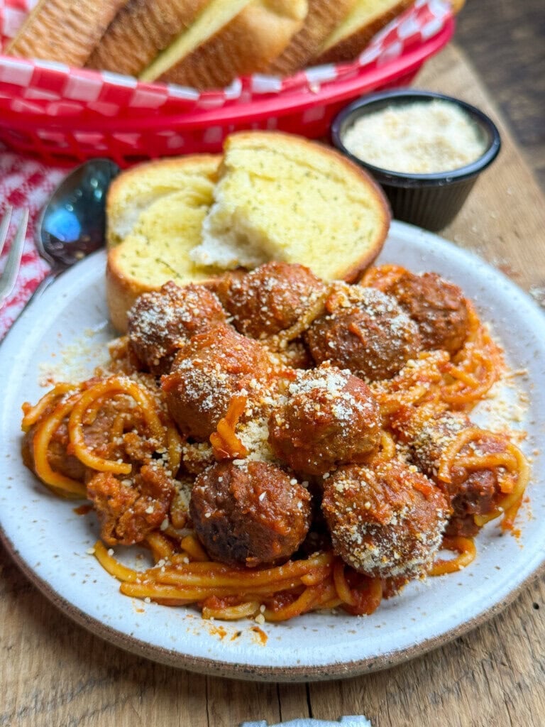 A plate of spaghetti topped with meatballs and grated cheese, served with garlic bread. A basket of breadsticks and a small bowl of grated cheese are in the background on a wooden table.