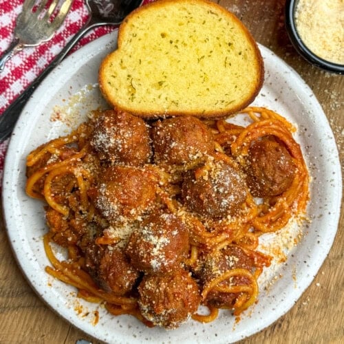 A plate of spaghetti with tomato sauce and meatballs, topped with grated cheese, served with a slice of garlic bread. A red and white napkin, a spoon, and a bowl of grated cheese are nearby.