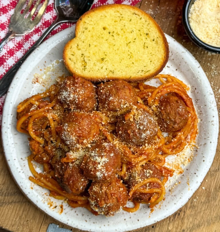 A plate of spaghetti with tomato sauce and meatballs, topped with grated cheese, served with a slice of garlic bread. A red and white napkin, a spoon, and a bowl of grated cheese are nearby.