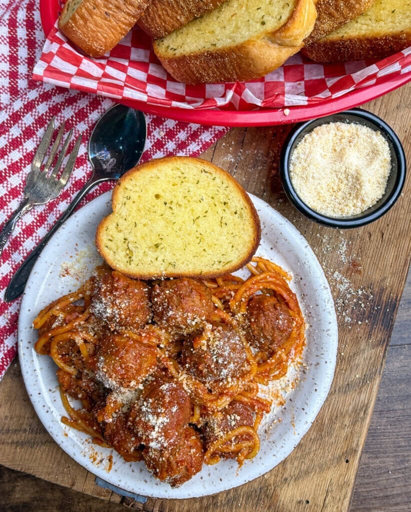A plate of spaghetti with meatballs topped with grated cheese, served with a slice of garlic bread. A basket of more garlic bread, a spoon and fork, and a small bowl of grated cheese are nearby on a wooden table.