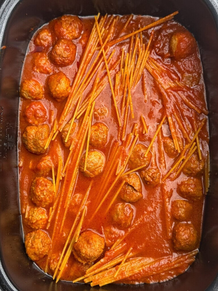 Meatballs and broken uncooked spaghetti noodles in tomato sauce are shown in a rectangular black cooking dish. The noodles and meatballs are partially submerged in the red sauce.