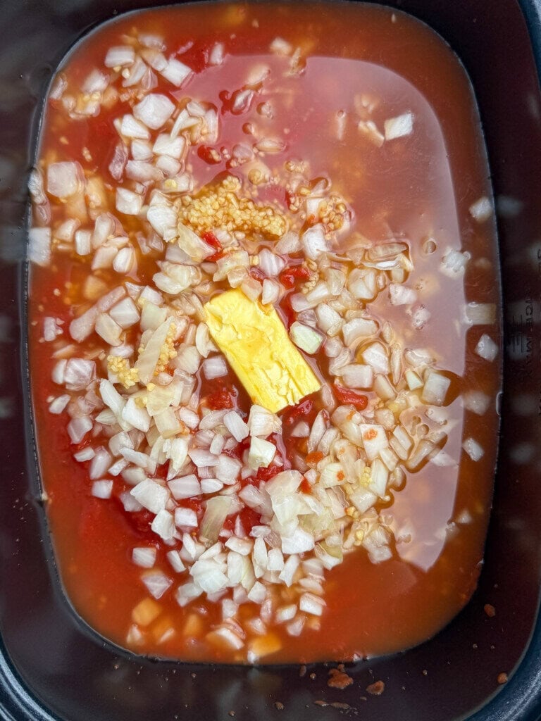 Chopped onions, minced garlic, a pat of butter, and tomato sauce or soup in a black slow cooker or pot, ready to be cooked.