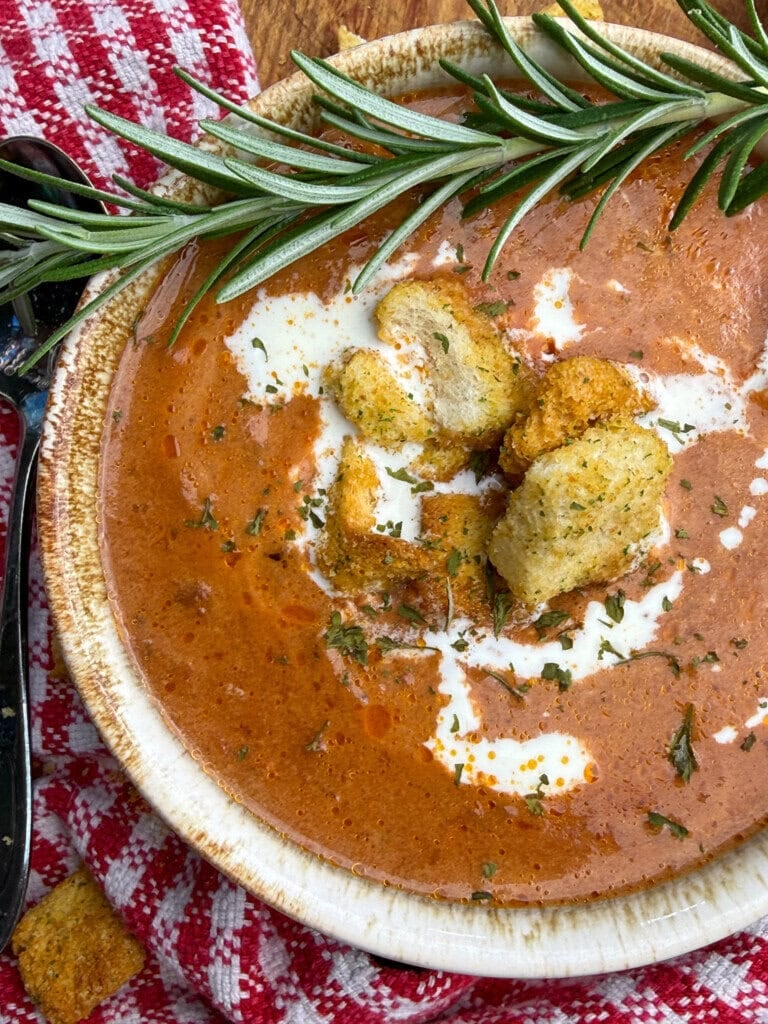 A bowl of creamy tomato soup topped with croutons and herbs, garnished with a sprig of rosemary. The bowl sits on a red and white checkered cloth, with a spoon nearby.