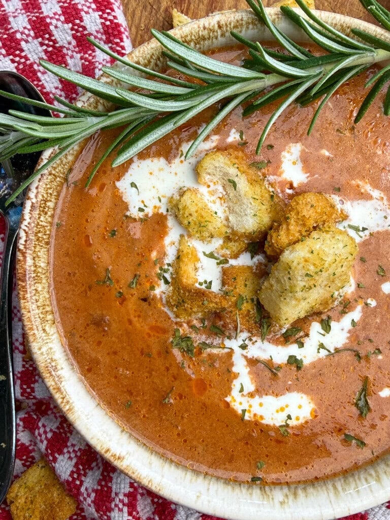 A bowl of creamy tomato soup garnished with croutons, herbs, a swirl of cream, and a sprig of rosemary, set on a red checkered cloth.