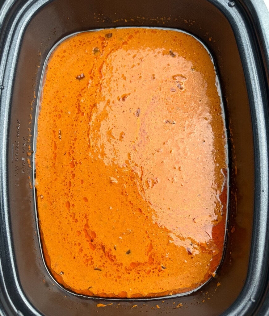 A slow cooker filled with creamy tomato soup and small specks of herbs and spices throughout.