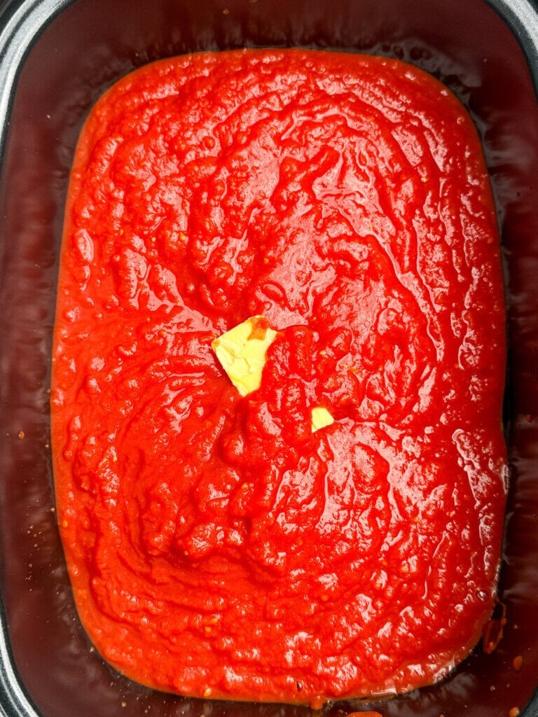 A close-up view of a rectangular pan filled with smooth, bright red crushed tomatoes, topped with a small piece of butter melting in the center.