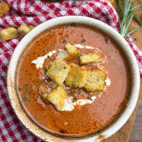 A bowl of tomato soup topped with croutons and a swirl of cream, placed on a wooden surface next to a red and white checkered cloth and a sprig of fresh rosemary.