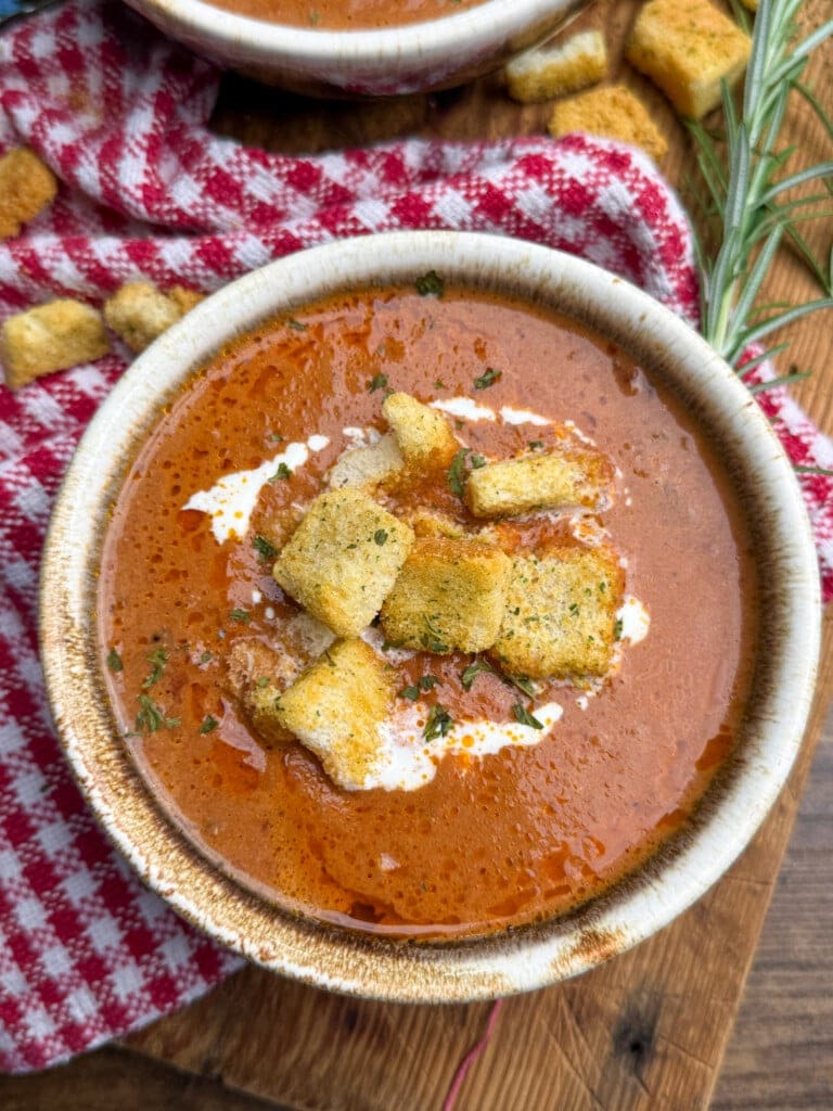 A bowl of tomato soup topped with croutons and a swirl of cream, placed on a wooden surface next to a red and white checkered cloth and a sprig of fresh rosemary.