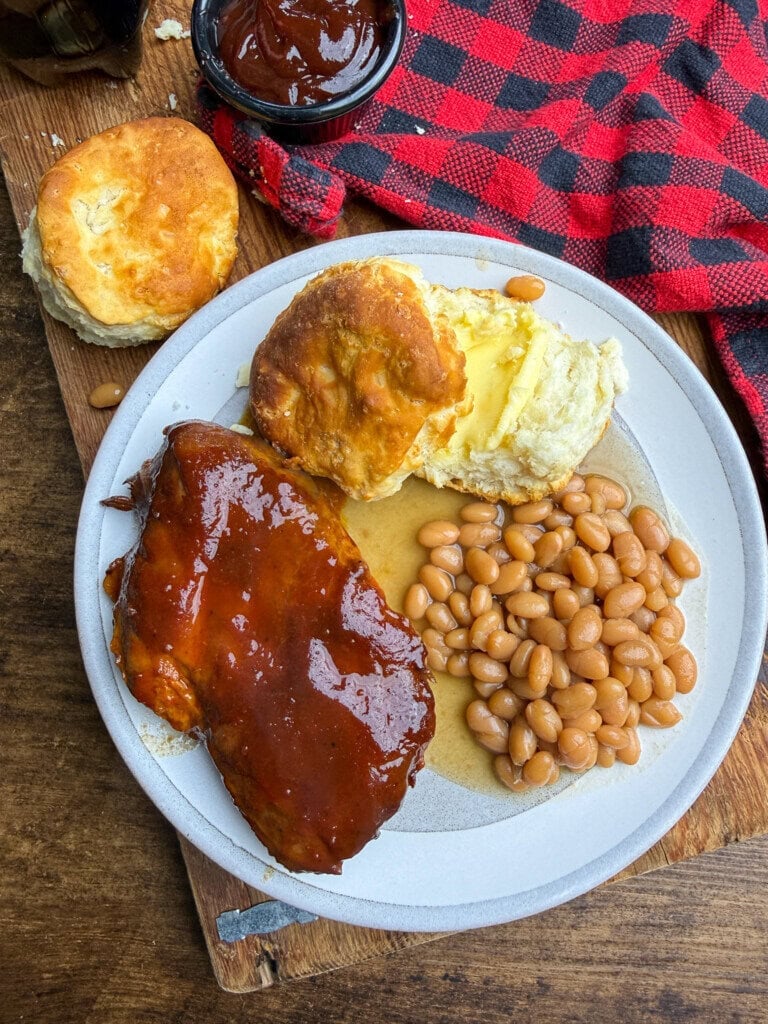 A plate of barbecue chicken with sauce, baked beans, and two buttery biscuits sits on a table next to a red plaid cloth, with extra biscuits and barbecue sauce nearby.