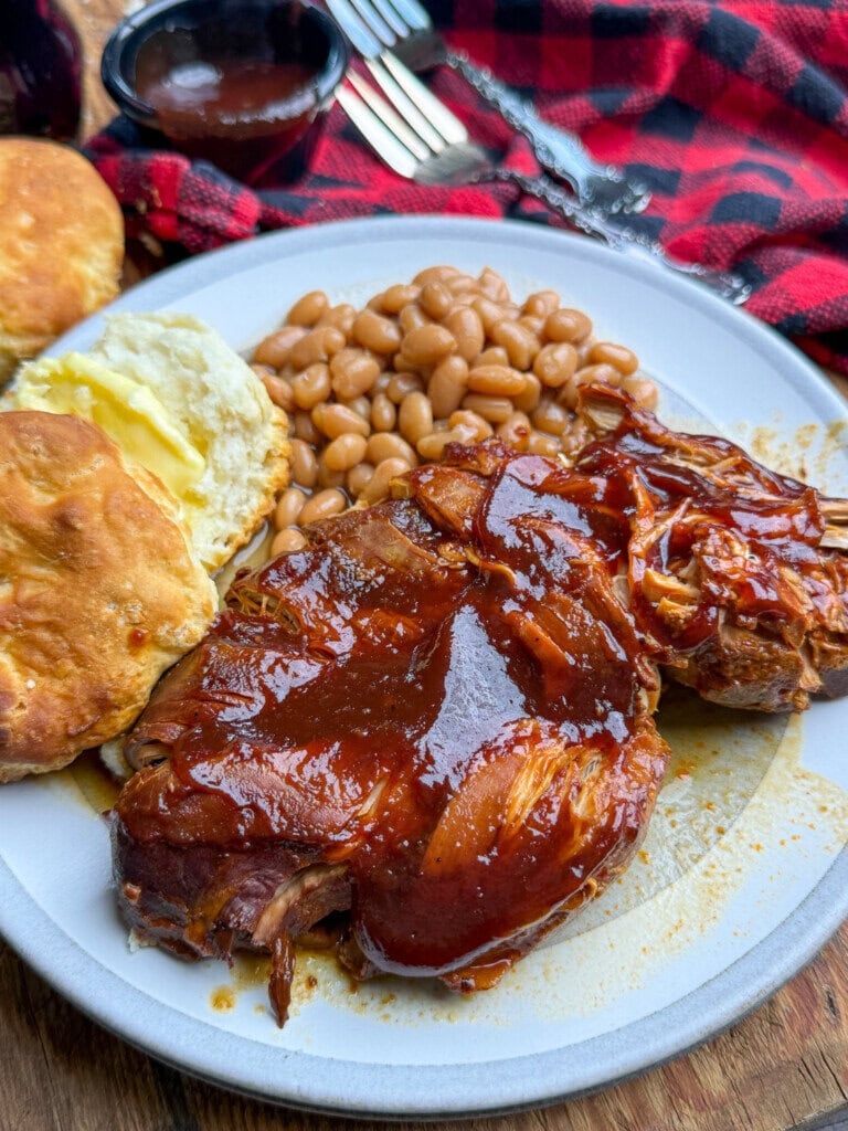 Plate with barbecue pulled chicken covered in sauce, baked beans, and a buttered biscuit. A red plaid cloth, fork, and cup of extra barbecue sauce are in the background.