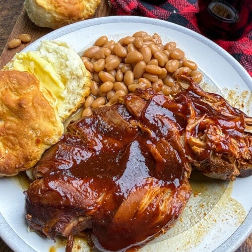 Plate with barbecued pulled chicken, baked beans, and a buttered biscuit. In the background are a cup of barbecue sauce, another biscuit, a red plaid napkin, and a bottle of Coca-Cola on a wooden table.