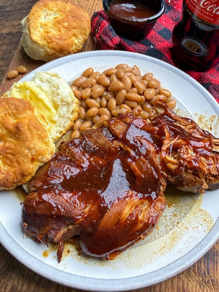Plate with barbecued pulled chicken, baked beans, and a buttered biscuit. In the background are a cup of barbecue sauce, another biscuit, a red plaid napkin, and a bottle of Coca-Cola on a wooden table.