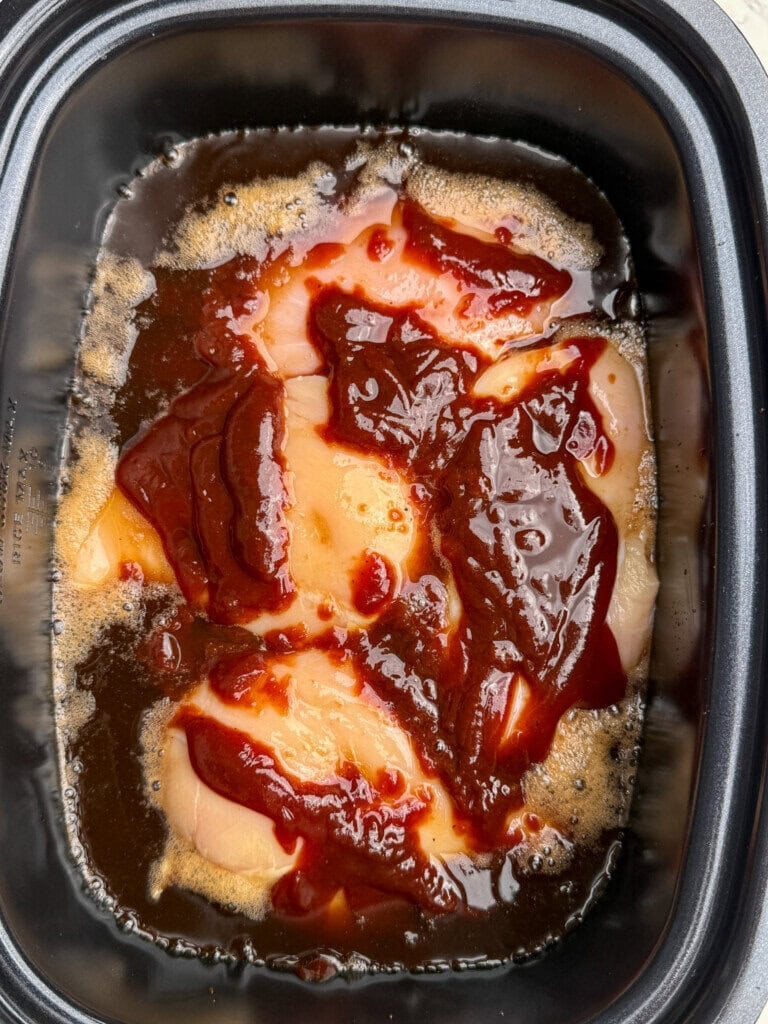 Raw chicken breasts topped with barbecue sauce are placed in a black slow cooker, ready to be cooked. The sauce is spread unevenly over the chicken, and some liquid is visible around the edges.