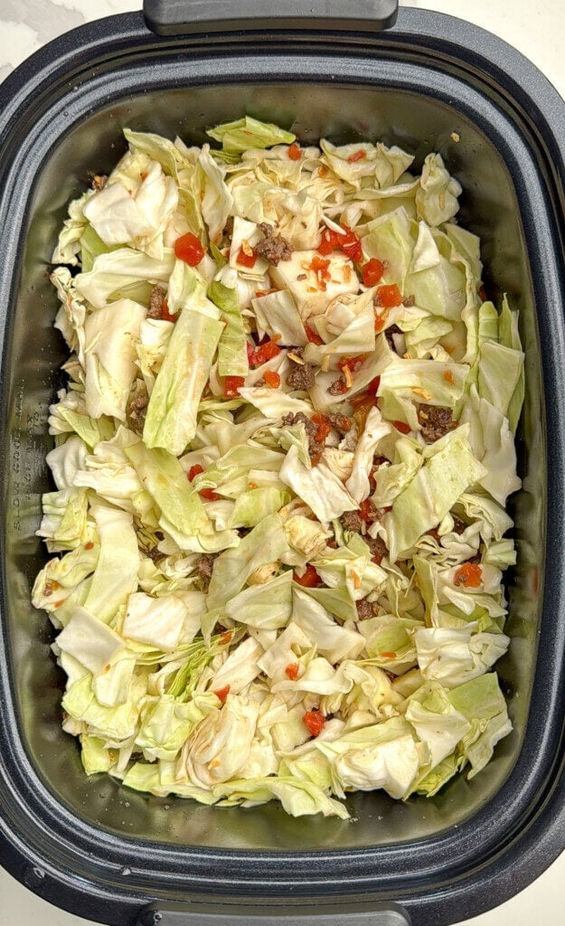 Chopped cabbage, ground meat, and small diced red tomatoes in a slow cooker, ready to be cooked.