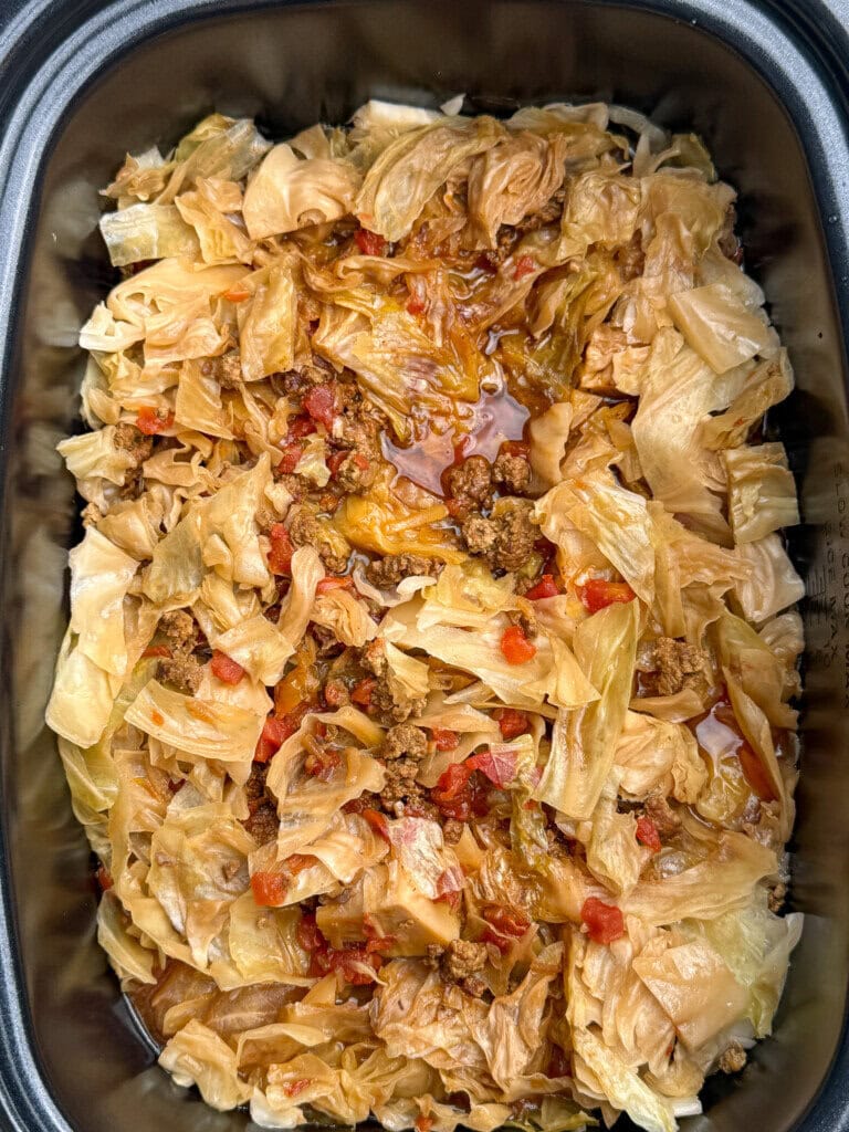 A close-up of cooked cabbage, ground meat, and diced tomatoes in a slow cooker, showing a savory, saucy dish with browned edges and visible seasonings.