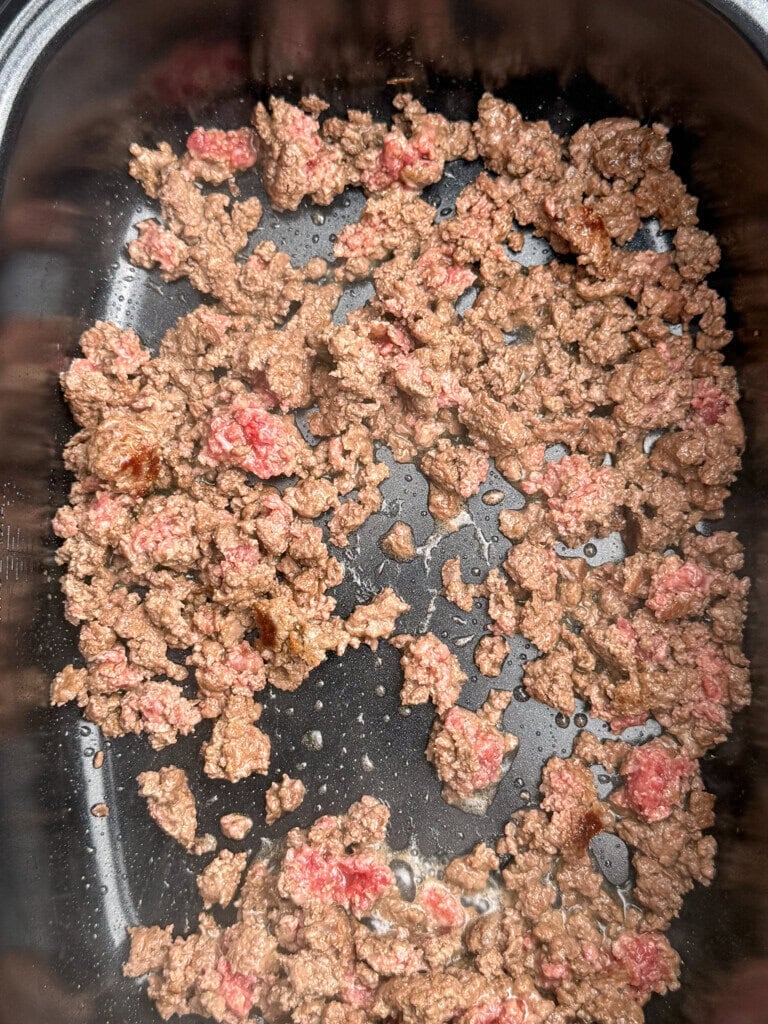 Ground beef cooking in a Crock pot, with some parts browned and others still pink, surrounded by a bit of fat and juices.