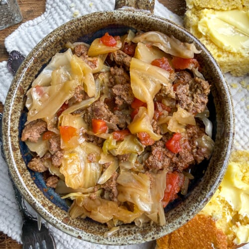 A rustic bowl filled with cooked ground beef, cabbage, and tomatoes sits on a textured napkin, with buttered cornbread pieces on the side. Fork and knife rest nearby.