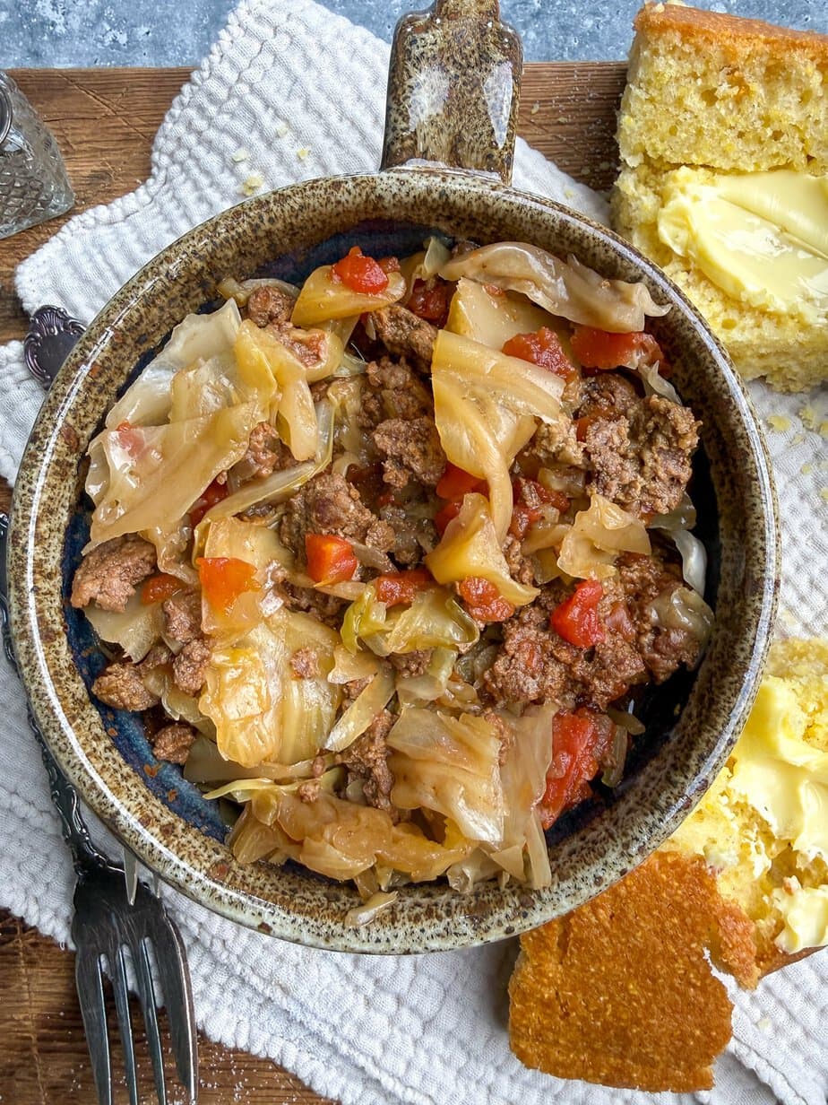 A rustic bowl filled with cooked ground beef, cabbage, and tomatoes sits on a textured napkin, with buttered cornbread pieces on the side. Fork and knife rest nearby.