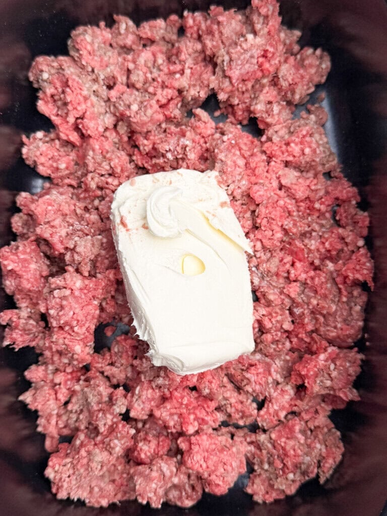 Raw ground beef spread in a pan with a block of cream cheese placed on top, ready to be cooked or mixed.