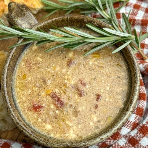 A bowl of creamy soup with visible herbs and bits of meat, garnished with a sprig of fresh rosemary, sits on a red checkered cloth next to pieces of bread.