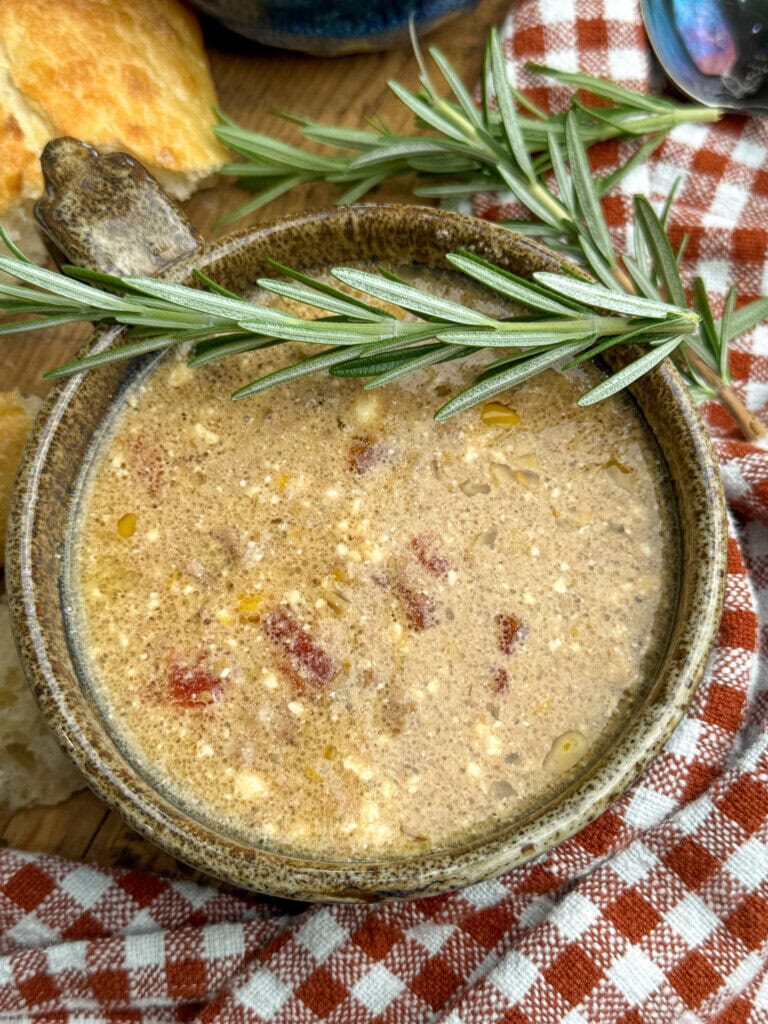 A bowl of creamy soup with visible herbs and bits of meat, garnished with a sprig of fresh rosemary, sits on a red checkered cloth next to pieces of bread.