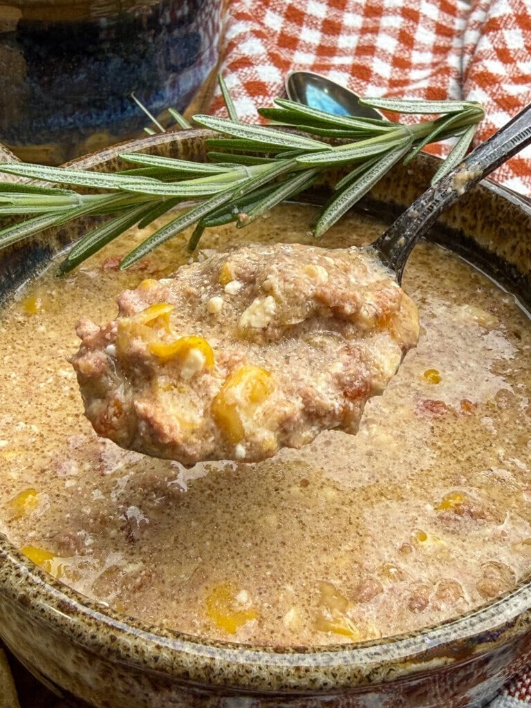 A close-up of a spoonful of hearty soup with ground beef, corn, and cheese, held over a rustic bowl. Fresh rosemary and a red checkered cloth are in the background.