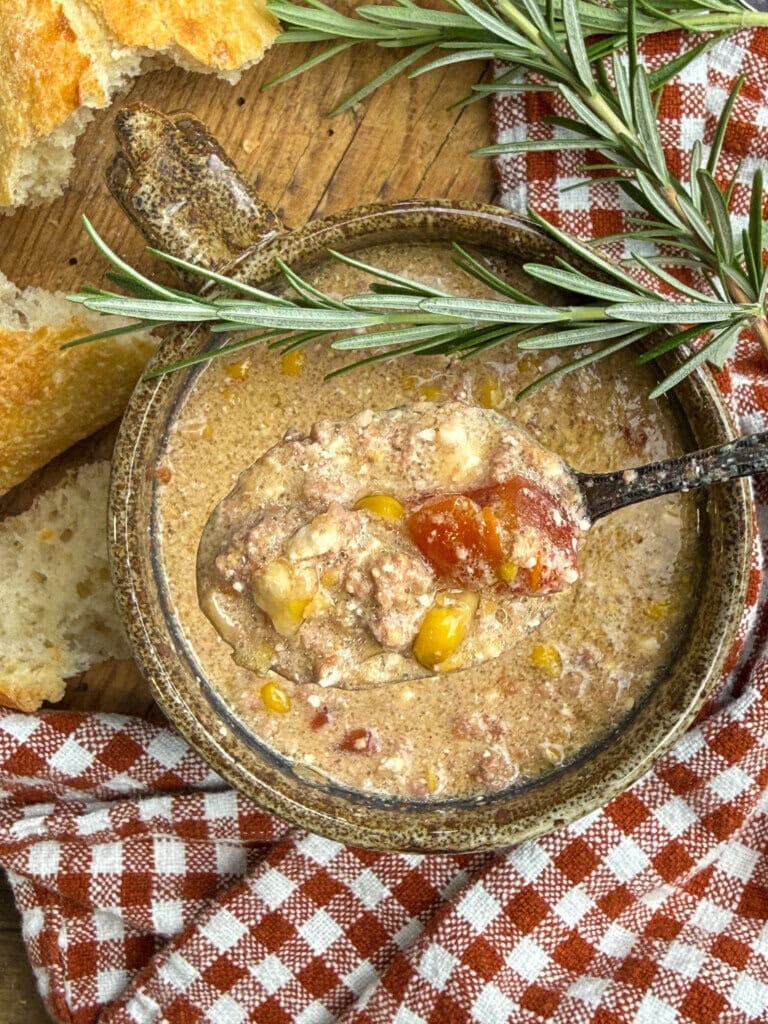 A bowl of creamy soup with ground meat, corn, and tomatoes, garnished with rosemary. Bread pieces and a red plaid cloth are nearby on a wooden surface.
