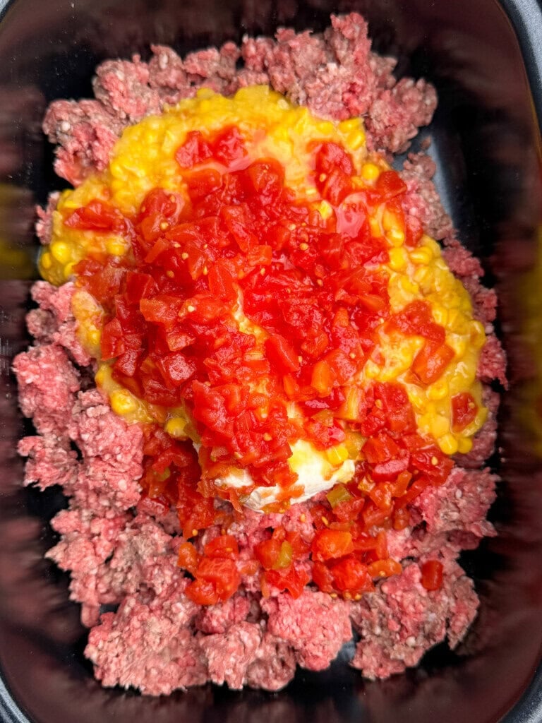 Uncooked ground beef topped with creamed corn, chopped tomatoes, and diced red peppers in a black container, ready to be mixed or cooked.