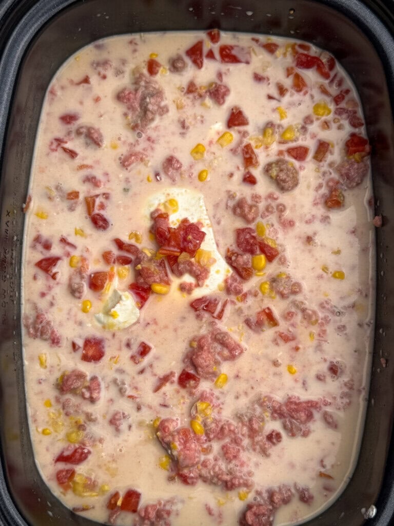 A slow cooker filled with a creamy mixture of ground beef, diced tomatoes, corn, and a block of cream cheese.