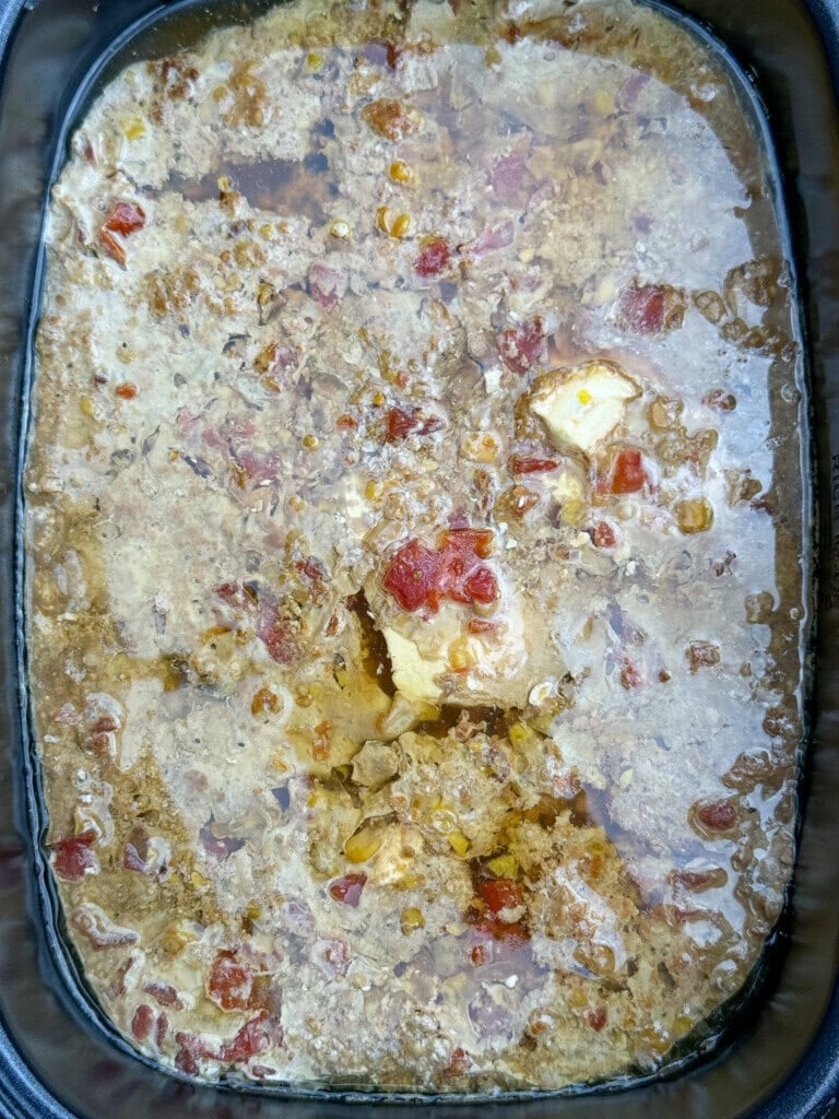 A close-up of a slow-cooked dish with chunks of meat, diced tomatoes, and visible oil in a black slow cooker.