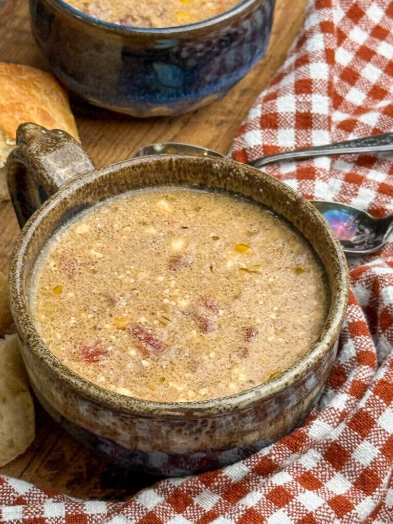 A rustic ceramic bowl filled with creamy soup, bits of meat and grains visible. It sits on a wooden table next to a red and white checkered cloth, a spoon, and a piece of crusty bread.
